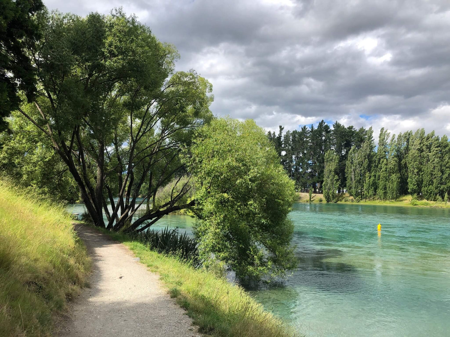 Gravel path along Hawea River under cloudy sky and trees