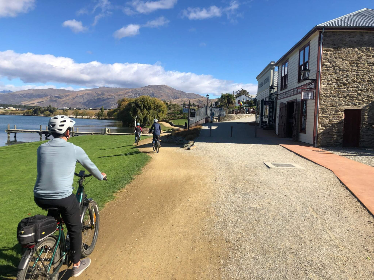 Cyclists riding e-bikes past heritage buildings along a lakefront trail in Bannockburn